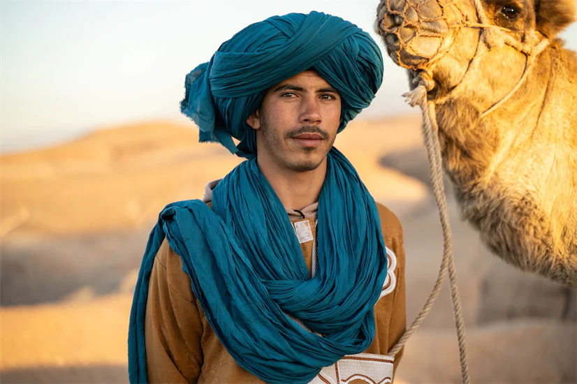 Local Berber guide in traditional blue turban standing beside a camel in the Moroccan desert at golden hour.